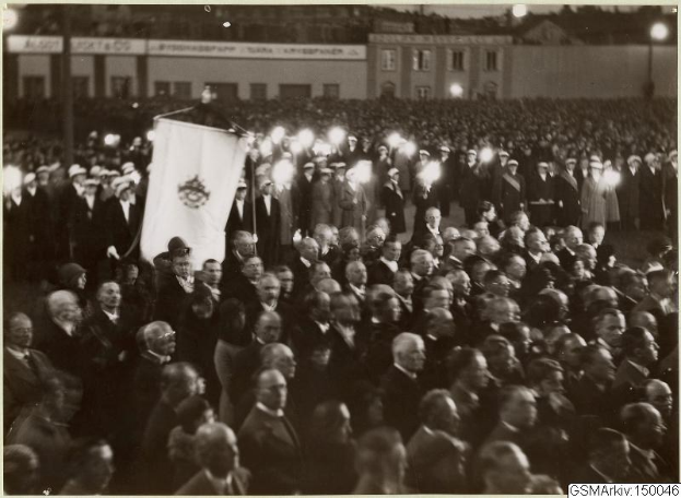 Schwarzes und weißes Foto einer großen Menge vor einem Gebäude, mit einer Person, die eine Fahne hält, Stangen und Lichter im Hintergrund und dem Text "Gustav Klimts Rede an der Berliner Mauer" unten.