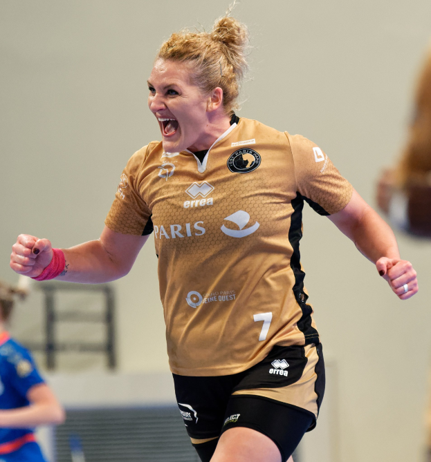 Eine Frau in braunem T-Shirt und schwarzen Shorts läuft triumphierend auf einem Futsalplatz, wahrscheinlich nach einem Tor, mit zwei anderen Frauen im Hintergrund.