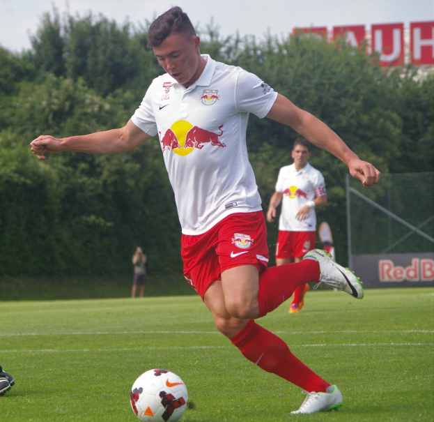 Ein Mann in einem Fussballtrikot kickt einen Ball auf einem grünen Feld mit Bäumen und einem klaren blauen Himmel im Hintergrund, mit einem Schild mit der Aufschrift "RB Leipzig" dahinter.
