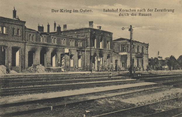 Schwarzes und weiße Foto des Bahnhofs Korschen in Ostern, Deutschland, mit Schienen, Gebäuden, Menschen, Bäumen und Himmel, mit Stationsnamen-Text oben und unten.