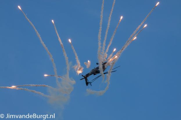 Ein Hubschrauber fliegt durch die Luft und hinterlässt eine Rauchspur, die eine Feuerlinie am Himmel bildet, mit Text in der linken unteren Ecke.