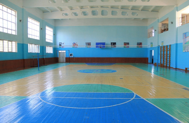 Großer Turnsaal mit blau-weißem Boden, Basketballfeldmarkierungen, einem Basketballkorb mit Netz, Stützpfosten, einer Tür, Fenstern, Wandplakaten, einem Geländer und einem Dach.