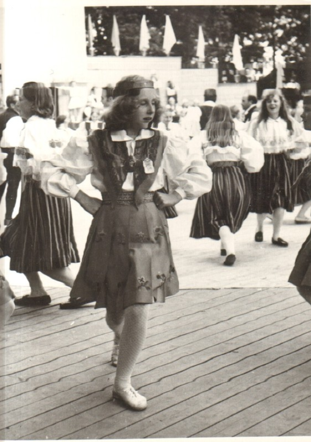 Eine Gruppe von Frauen in traditioneller bayrischer Tracht tanzt auf einem Holzboden, mit einer Wand, Bäumen und anderen Gegenständen im Hintergrund; Text ist unten im Bild sichtbar.