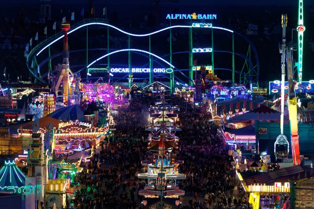 Eine große Menschenmenge versammelte sich um einen hell erleuchteten Karneval in der Nacht, umgeben von Vergnügungsgeräten, Polen, Fahnen und Namensschildern, mit einem dunklen Hintergrund.