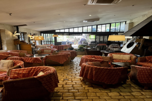 Eine geräumige Hotellobby mit zahlreichen Stühlen, Tischen, Sofas mit Kissen, Wandlampen, einer Deckenklimaanlage und großen Glasfenstern an der Rückseite.