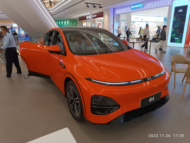 Oranges Elektroauto in einer Ausstellungshalle umgeben von Menschen, Stühlen, einem Wagen und Ladenschildern im Hintergrund.