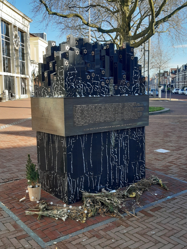 Denkmal in einem Stadtplatz für Holocaust-Opfer mit einem Baum davor, einem Topfpflanze auf dem Boden, verstreuten trockenen Blättern und Gebäuden, Fahrzeugen, Masten und Bäumen im Hintergrund.