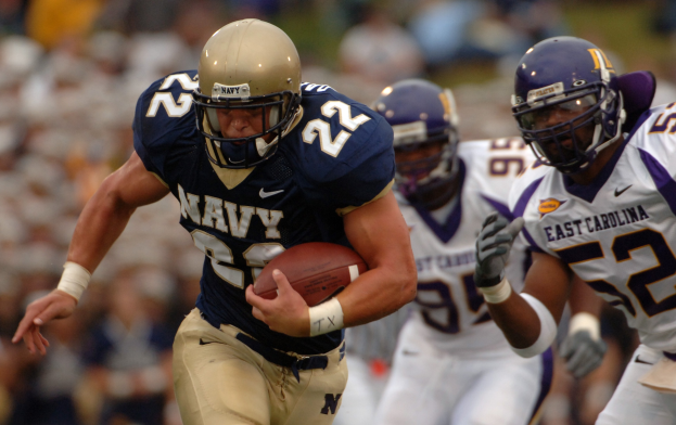 Ein Football-Spieler in einem marineblauen Trikot und Helm rennt mit dem Ball während eines Spiels, der Hintergrund durch ihre Geschwindigkeit unscharf.