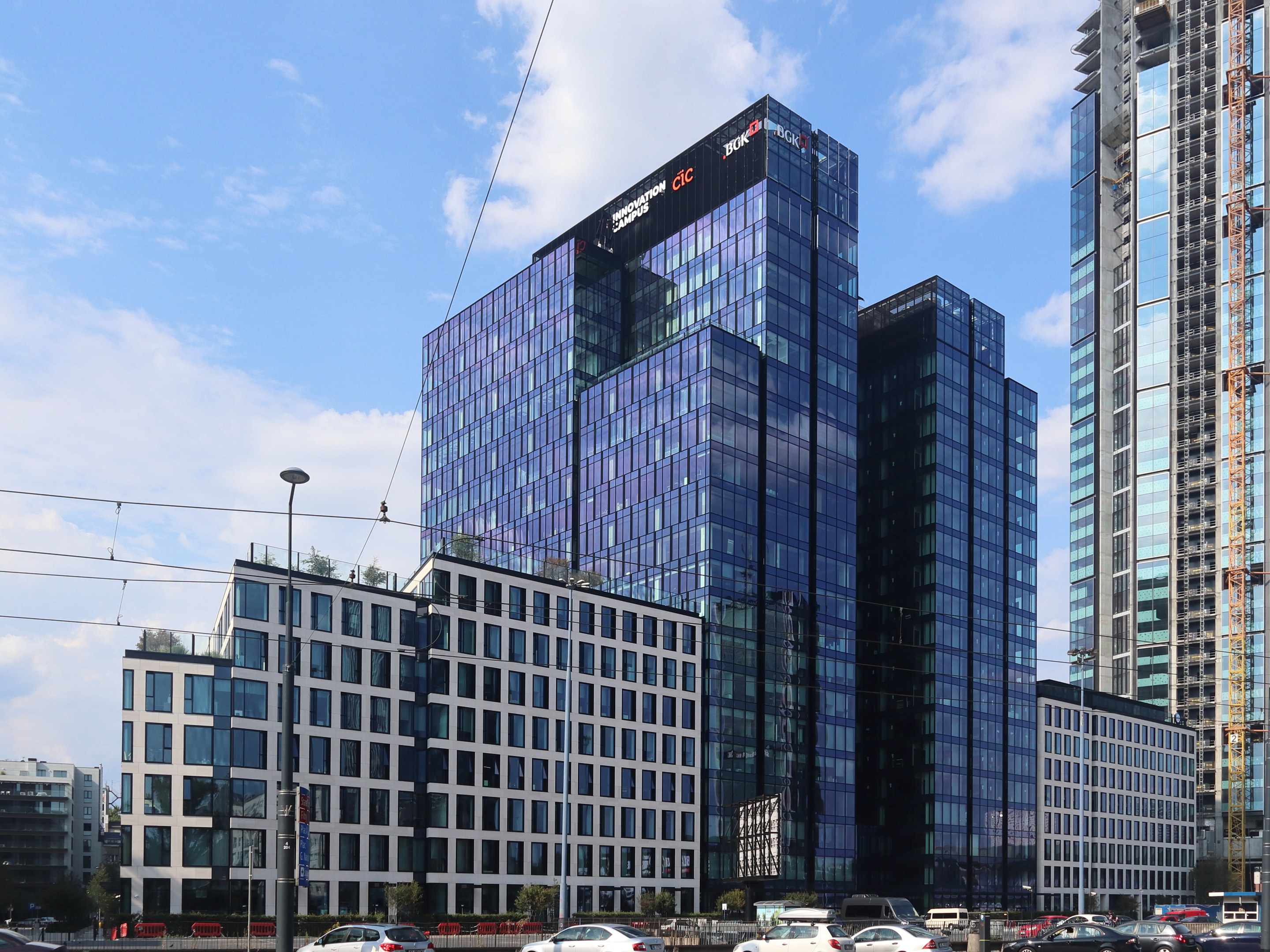 Außenansicht des Deutschen Bank Hauptsitzes in Frankfurt, Deutschland, mit einem mehrstöckigen Gebäude mit zahlreichen Fenstern, umgeben von städtischer Infrastruktur wie Straßenlaternen, Fahrzeugen, Bäumen und einem bewölkten Himmel.