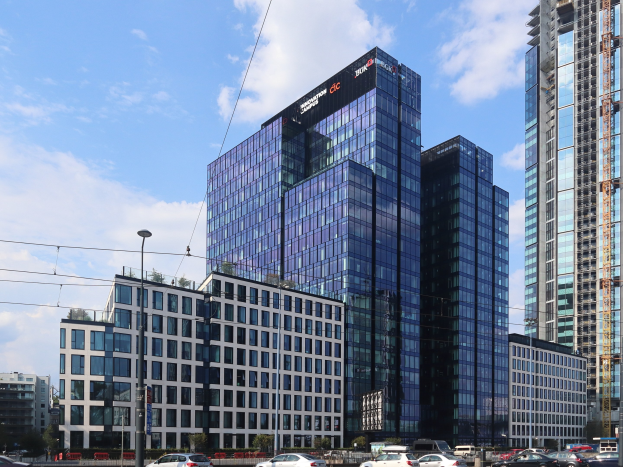 Außenansicht des Deutschen Bank Hauptsitzes in Frankfurt, Deutschland, mit einem mehrstöckigen Gebäude mit zahlreichen Fenstern, umgeben von städtischer Infrastruktur wie Straßenlaternen, Fahrzeugen, Bäumen und einem bewölkten Himmel.
