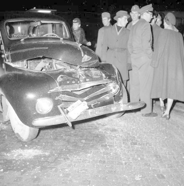 Schwarzes Und Weißes Bild Eines Schwer Geschädigten Autos Am Straßenrand Mit Einem Zerdrückten Kühlergrill Und Zerbrochenen Fenstern, Umgeben Von Menschen, Die Die Szenerie Begutachten.