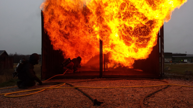 Feuerwehrleute löschen ein Gebäudebrand, während ein Rohr vor ihnen auf dem Boden liegt, mit Schuppen, Bäumen und einem klaren blauen Himmel im Hintergrund.