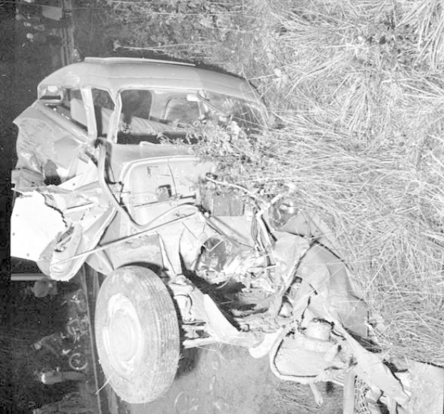 Schwarz-weiß-Foto eines stark beschädigten Autos mit eingedrückter Front und zersplitterten Fenstern am Straßenrand, mit einem Baum und verstreuten Gegenständen im Hintergrund.