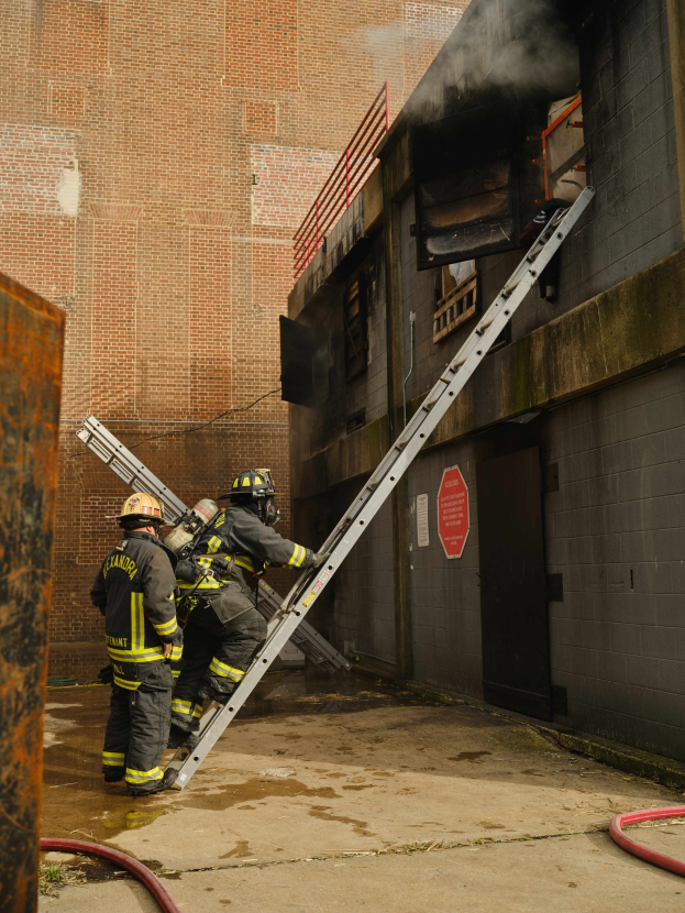 Feuerwehrleute in Helmen klettern eine Leiter zu einem raucherfüllten Gebäude hinauf, mit einem Metallobjekt links und Rohren am Boden.