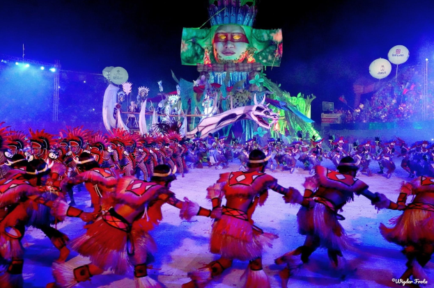 Eine Gruppe von Menschen in bunten Kostümen tanzt auf einer Bühne vor einem sitzenden Publikum, mit Lichtern, Pfählen, Ballons und einem Bildschirm, der eine Person in den Hintergrund zeigt, unter einem sichtbaren Himmel, bei der Rio-Karneval 2014.