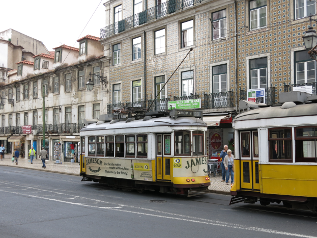 Zwei gelbe und weiße Straßenbahnen fahren eine Straße mit hohen Gebäuden entlang, mit Menschen auf dem Gehweg, Laternen und Texttafeln zu sehen, unter einem klaren Himmel.