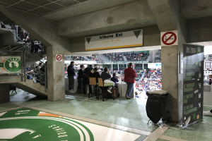 Gruppe von Menschen, die vor einer sitzenden Menge in einem Stadion stehen, mit Schildern, die "Boston Celtics vs New York Jets" lesen, einem Müllcontainer, einer Tafel mit Text, einer Treppe mit Geländern und einem Dach mit Deckenlampen.
