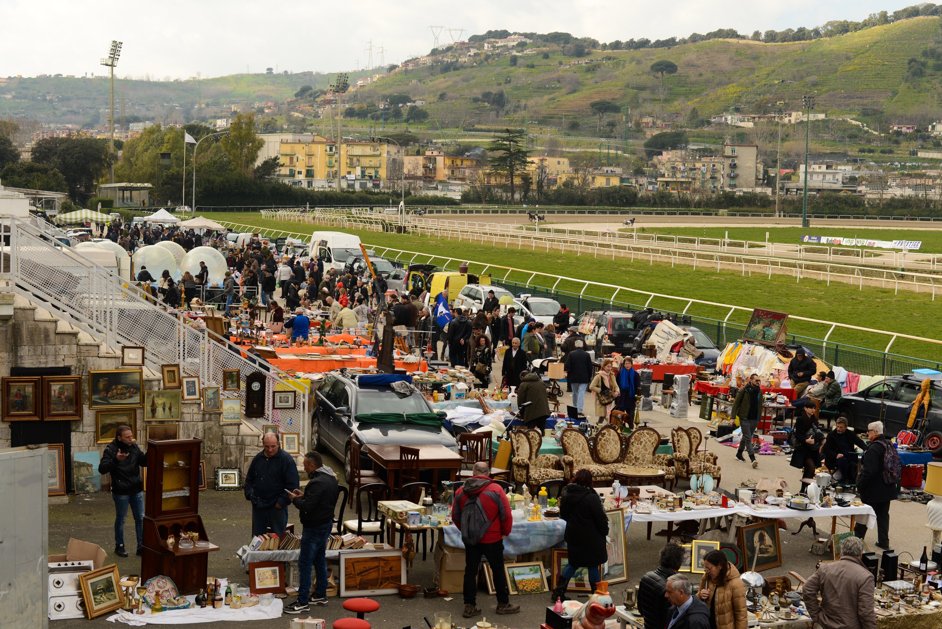 Eine große Gruppe von Menschen auf einem Flohmarkt mit Tischen, auf denen Gegenstände wie Foto Rahmen und Stühle ausgelegt sind, Fahrzeuge in der Nähe geparkt, Geländer, Stufen, Bäume, Gebäude, Laternenpfähle, Hügel und ein bewölkter Himmel im Hintergrund.
