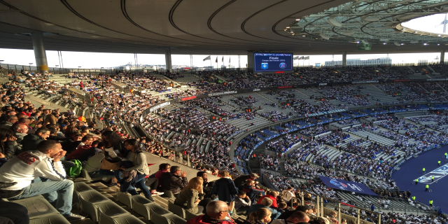 Eine große Menschenmenge sitzt im Allianz Stadion in München, Deutschland, und schaut ein Fussballspiel. Auf der rechten Seite befindet sich eine Bühne mit ein paar Leuten darauf und im Hintergrund sind Fahnen, Stangen und ein Bildschirm zu sehen. Der Himmel ist oben im Bild zu sehen und das Stadion scheint das Allianz Stadion in München, Deutschland zu sein.
