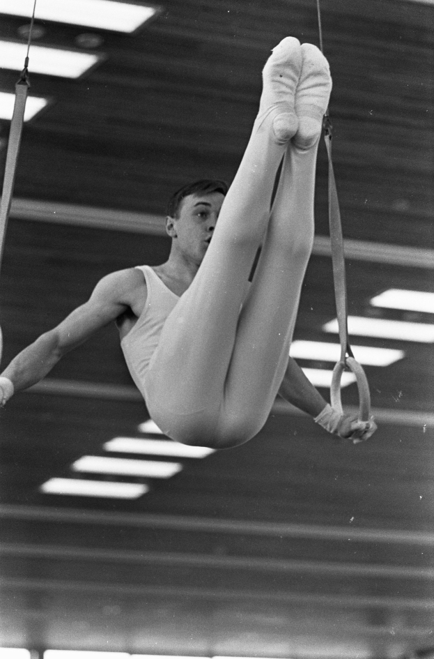 Ein Mann in der Luft, der Gymnastik an den Ringen in einem Fitnessstudio ausführt, beleuchtet von Deckenlampen.
