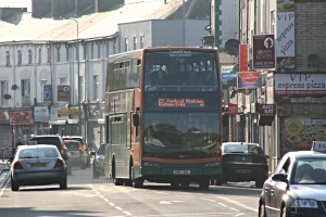 Eine Straße mit Autos und einem Bus vor Gebäuden mit Wänden, Fenstern, Tellern und Dächern, mit Plakaten und Bannern an den Wänden und einem Pfahl mit einer Straßenlaterne.