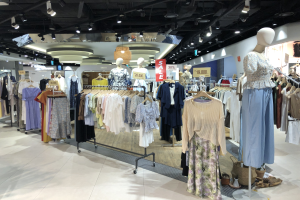 Bekleidungsgeschäft mit Schaufensterpuppen in Kleidern auf Bügeln, Texttafeln, Deckenbeleuchtung und diversen Bodenartikeln.