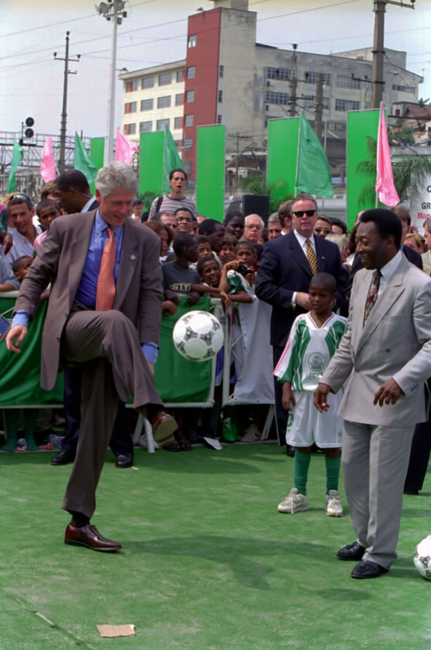 Ein Mann in einem Anzug, der einen Fußball auf einem Feld mit Zuschauern, darunter Menschen mit Fahnen und Kinder an einem Zaun, sowie Gebäuden und einem bewölkten Himmel im Hintergrund kickt.