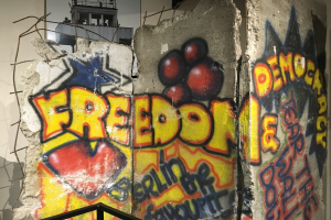 Eine Wand mit Graffiti, das "Freiheit und Demokratie" in großen, schwarzen Buchstaben schreibt, ein Geländer unten und ein Gebäude mit Fenstern im Hintergrund.