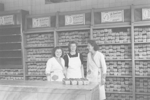 Drei Frauen in Schürzen lächeln vor einem Bäckertisch mit Lebensmitteln, mit Regalen voller Lebensmittel und Wandtafeln im Hintergrund, in Schwarz-Weiß.