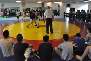 Zwei Menschen ringen auf einer Matte, ein Mann in einem weißen T-Shirt steht in der Nähe; Zuschauer sehen von den Seiten und dem Sitzbereich zu, mit Spiegeln, Trittbägen und Postern im Hintergrund.
