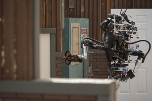 Ein humanoider Roboter mit ausgestreckten Armen und geneigtem Kopf steht vor einem Gebäude, seine großen runden Augen und der leicht geöffnete Mund deuten darauf hin, dass er sprechen könnte.