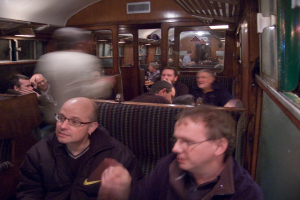 Zwei Männer sitzen auf einem Sitz in einem Zug, mit einer Gruppe von Menschen hinter ihnen, einem Eingang zwischen Abteilen oben und einem Fenster rechts.