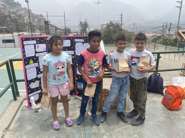 Eine Gruppe von Kindern, die auf einem Basketballfeld stehen und Papiertüten halten, mit Tüten und Gegenständen rechts daneben und einer Tafel mit Text links daneben, vor einem Hintergrund aus Zäunen, Pfosten, Straßenlaternen, Bäumen, Gebäuden, Hügeln und dem Himmel.