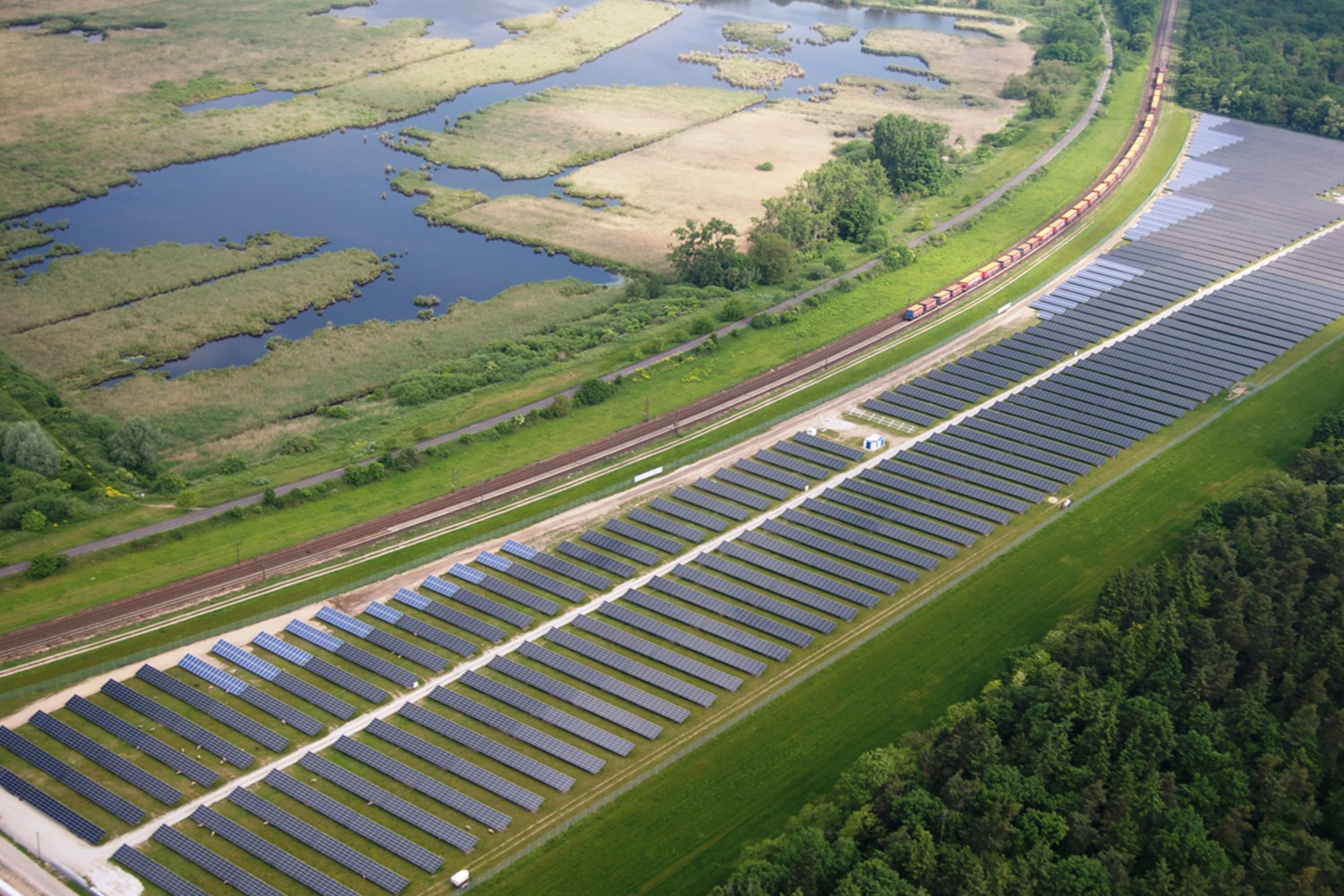Luftaufnahme einer Solar-Farm mit Solarpanelen, umgeben von Bäumen, Gras, Pflanzen und Wasser, mit einem Zug auf einer nahen Bahnstrecke.