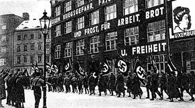 Schwarzes und wei├čes Foto einer Gruppe von Menschen, die mit Fahnen und Transparenten die Stra├če entlangmarschieren, mit einem Laternenpfahl links und einer Tafel mit der Aufschrift "Deutsche Nationalsozialistische Arbeiterpartei in Berlin" rechts.