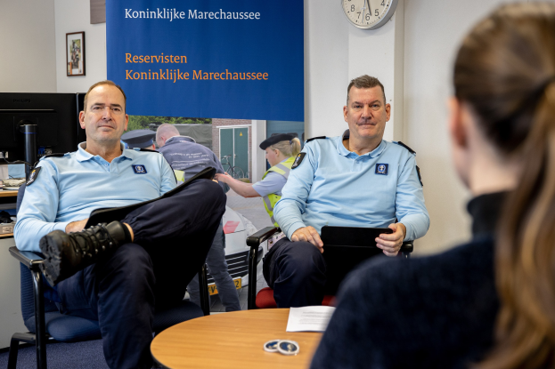 Zwei Polizeibeamte sitzen an einem Tisch mit einer Frau, umgeben von Dokumenten und Gegenständen, mit einem wandmontierten Banner, einer Uhr und Fotorahmen im Hintergrund und einem weiteren Tisch mit einem Monitor und Peripheriegeräten links daneben.