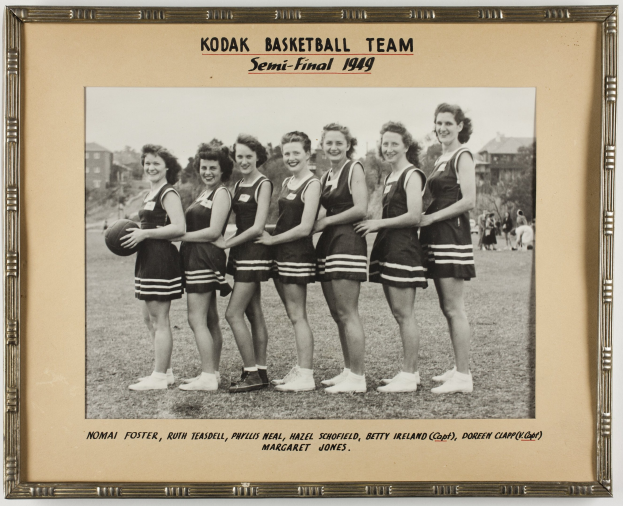 Schwarzes und weißes Foto einer Gruppe von Frauen der Kodak Basketball Mannschaft aus dem Jahr 1949, die lächeln und einen Basketball halten, mit Zuschauern, Bäumen, Gebäuden und Himmel im Hintergrund.