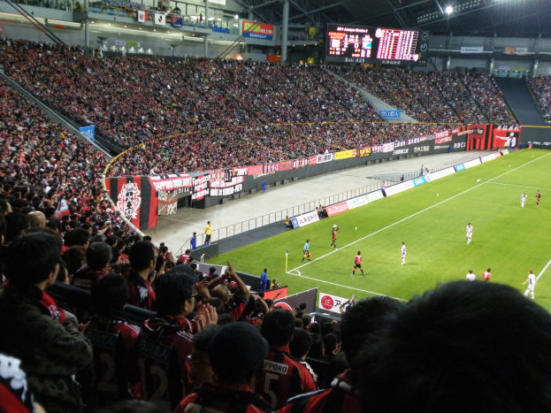 Ein Fußballspiel in einem großen Stadion mit Spielern auf dem Feld und Zuschauern in den Rängen, mit Bannern, einem Zaun, einem Anzeigebildschirm, Metallpfosten und Deckenleuchten.