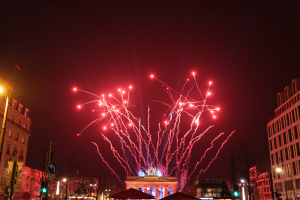 Eine Straßenansicht in Berlin am Silvesterabend, mit Gebäuden, Bäumen, Laternenpfählen, Verkehrszeichen, Zelten und Menschen, mit einem Feuerwerk-himmel im Hintergrund.