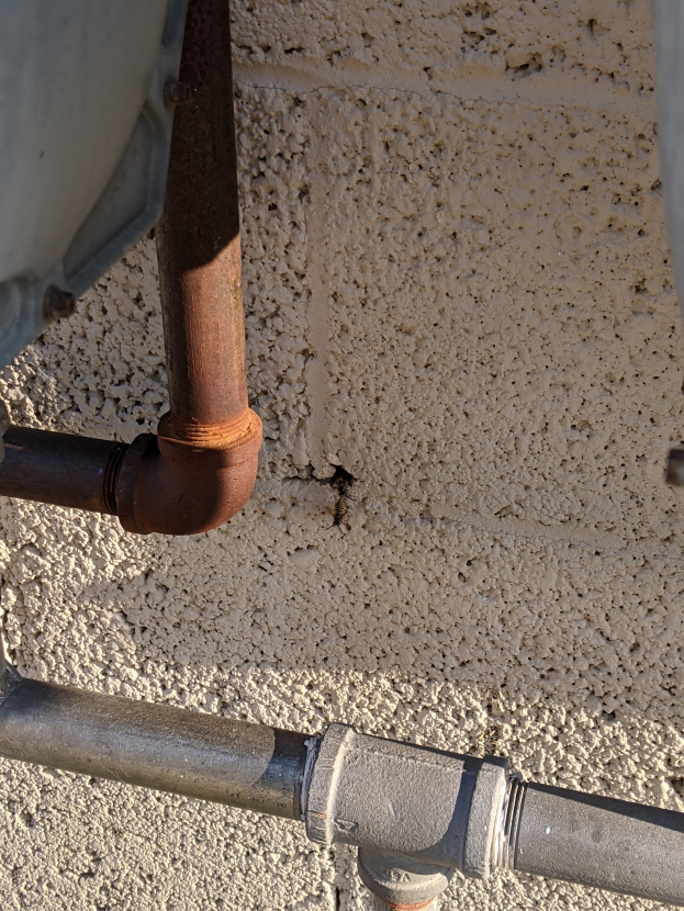 Ein Close-up eines Metallrohrs, das an einen Metallwasserheizer an der Seite eines Gebäudes angeschlossen ist, mit einer Wand im Hintergrund.