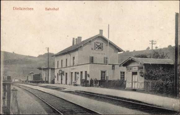 Schwarze und weiße Fotografie eines Bahnhofs in Dielkirchen, Deutschland, mit einem Zug auf den Gleisen, umgeben von Häusern, Strommasten, Bäumen, Menschen und Bergen sowie einem klaren Himmel im Hintergrund.
