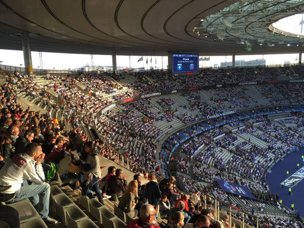 Großes Publikum in einem Stadion bei einem Förderball mit einer Bühne, Fahnen, Masten, einem Bildschirm und der Allianz Arena in München, Deutschland im Hintergrund.