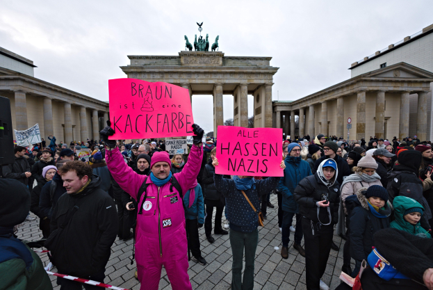 Eine Gruppe von Menschen steht vor dem Reichstagsgebäude in Berlin und hält Protestschilder gegen die anti-nazistische Bewegung der deutschen Regierung, mit Gebäuden und Statuen im Hintergrund unter einem klaren blauen Himmel.