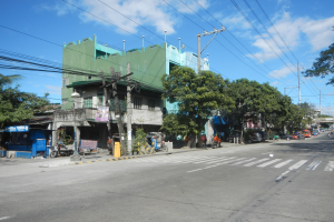 Eine Stadtstraße mit einem grünen Gebäude, fahrenden Fahrzeugen, Fußgängern auf dem Gehweg und Strommasten mit Drähten vor einem bewölkten Himmel.