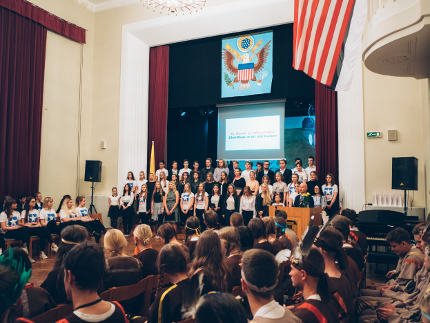Eine Gruppe von Menschen steht vor einem sitzenden Publikum bei einem Event, mit einem Podium, Mikrofonen, Flaggen, Lautsprechern, einem Bildschirm mit Text und einem Bild sowie prächtigen Vorhängen und einem Kronleuchter im Hintergrund.