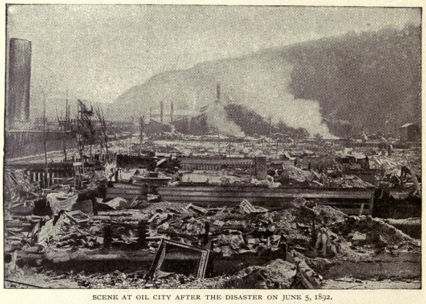 Schwarzes und weißes Foto einer Stadt nach einer Katastrophe am 5. Juni 1892, das Gebäude, Pfähle, Rauch und einen Hügel im Hintergrund zeigt, mit Text unten.