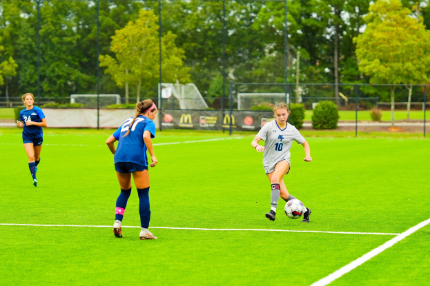 Gruppe junger Frauen beim Fußballspielen auf einem Rasenfeld mit Bäumen, Zäunen und einem Netz im Hintergrund.