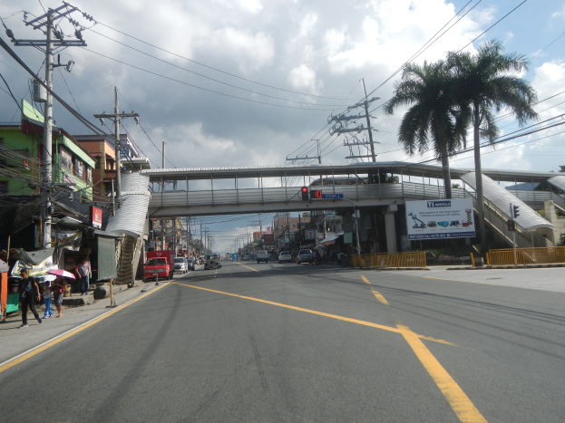 Stadtstraße mit Fahrzeugen, Fußgängern, Gebäuden, Bäumen, Brücke, Ampel, Strommasten und bewölktem Himmel.