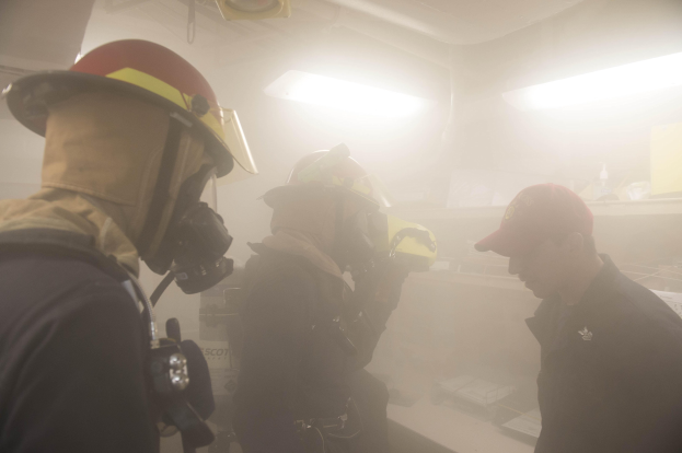 Feuerwehrleute mit Helmen und Rauch aus dem Mund, einer hält eine Kamera, während einer Trainingssitzung mit Hintergrundbeleuchtung und Gegenständen.