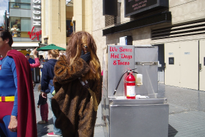 Eine Menschenmenge, darunter einige in Kostümen, versammelte sich vor einem Gebäude mit sichtbarer Beschriftung und Türen, mit einer Kiste und einem Feuerlöscher in der Nähe.